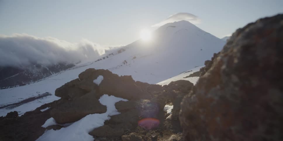 The sun sits just over the slope of a snow covered volcanic peak