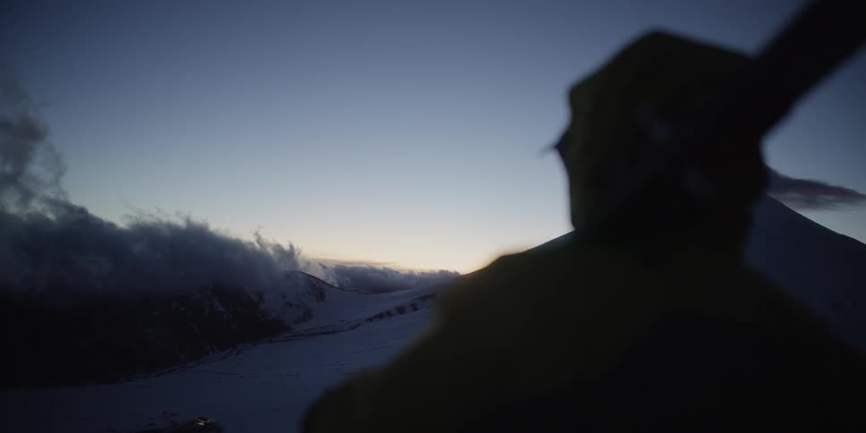 A skier looks out over snow covered mountains at twilight