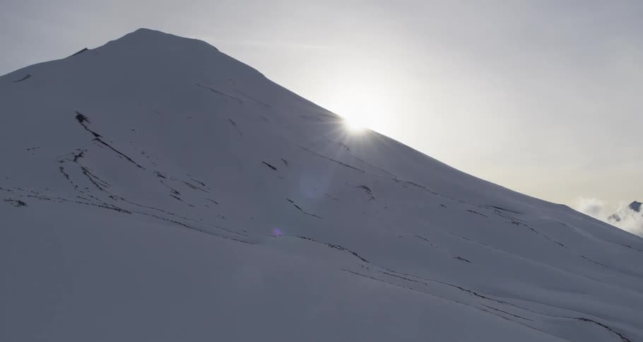 Aerial of the sun appearing behind a snowy volcano