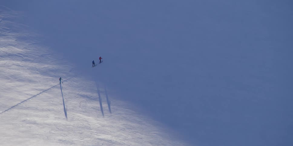 Three skiers move slowly across a shadowy mountain slope