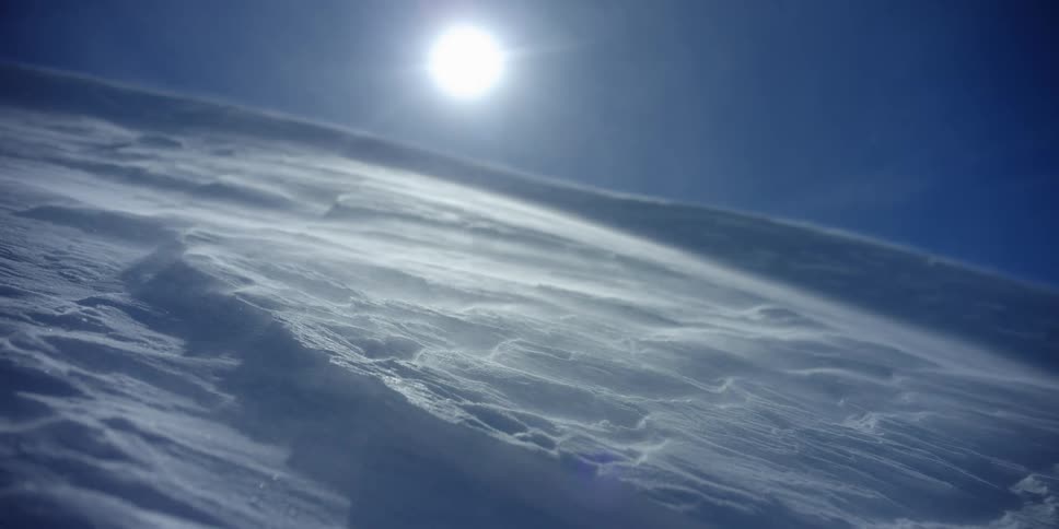 Drifting snow blows down a slope covered in snow under a bright sun