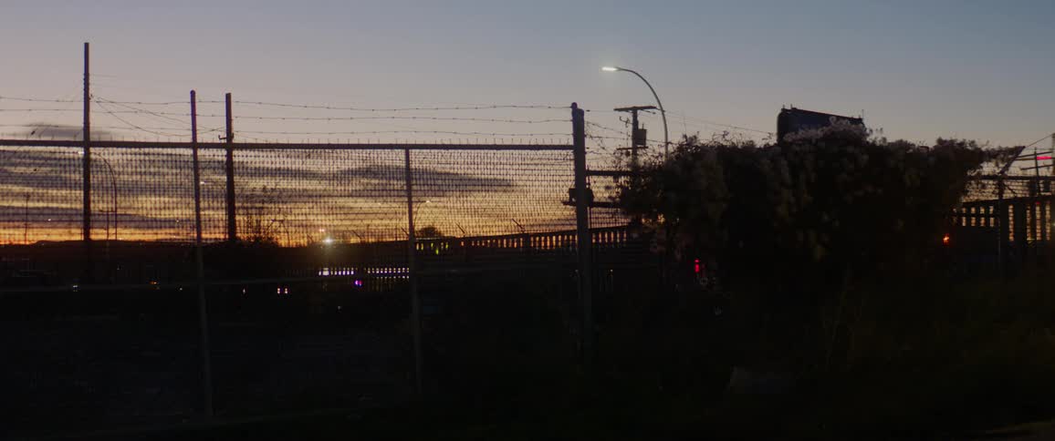 The headlights from passing cars and trucks on the road is visible through the fence and bushes