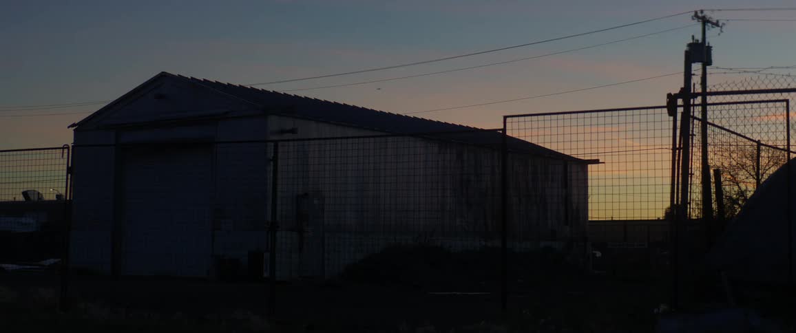 Building with large garage door stands on dark lot behind chain link fence backed by orange sky