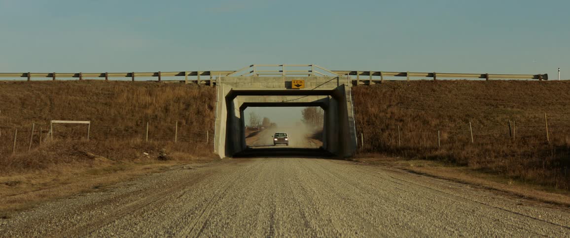 Vehicles drive down an overpass above a gravel road
