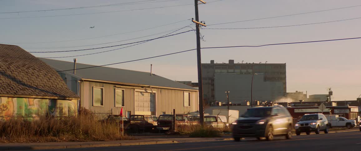 Cars travel on a road past the parking lot and buildings of a warehouse or factory