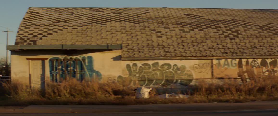 Car drive along road past graffiti strewn building on overgrown lot