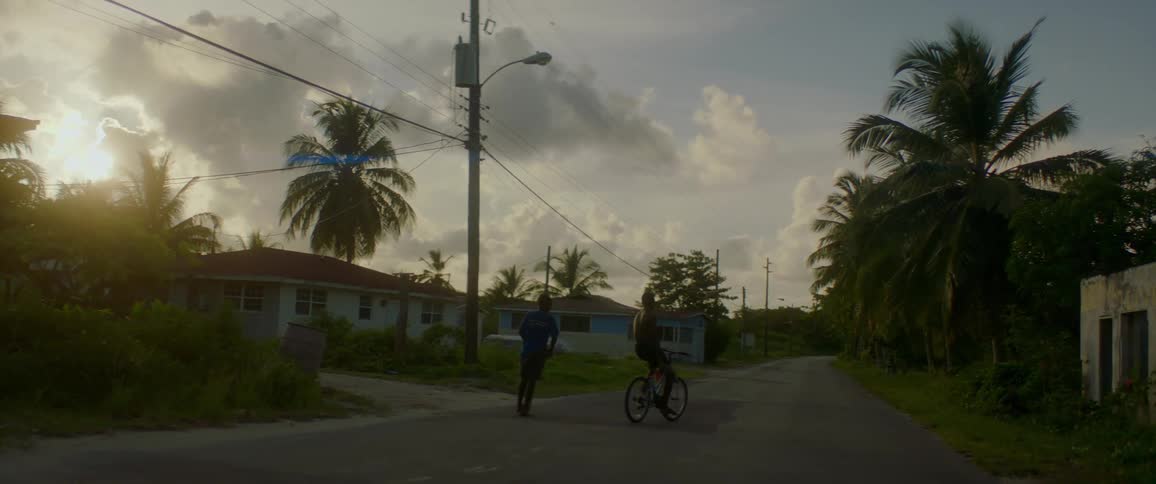Two black teens ride a bike and a scooter in a neighborhood street