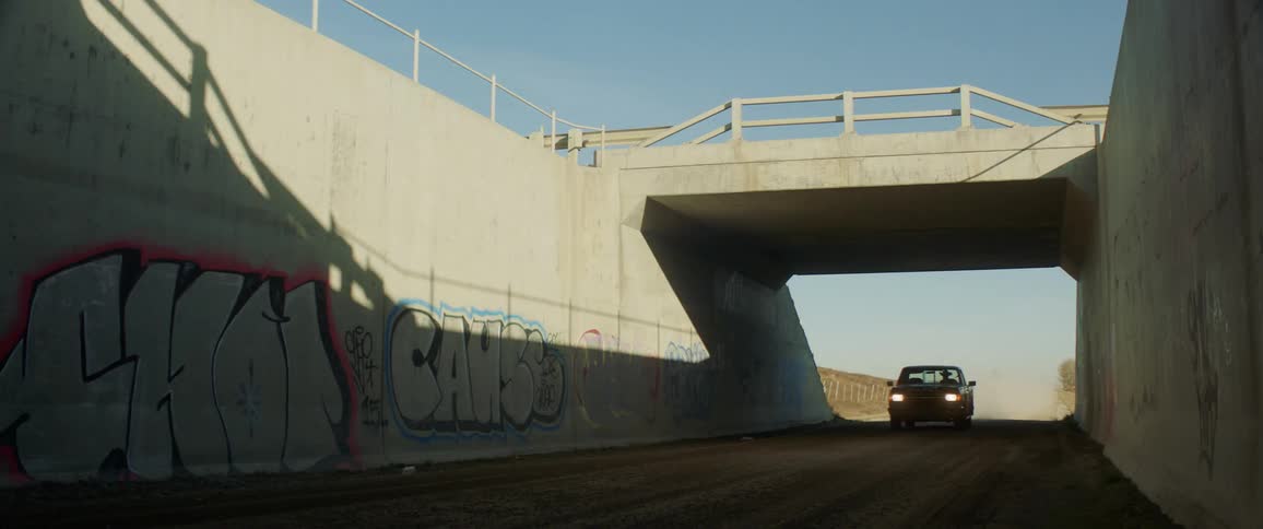 Older model pickup drives on dirt road under overpass