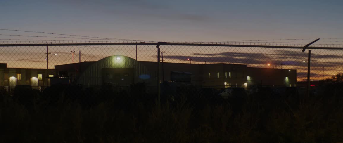 Large warehouse building and barbed wire fence silhouetted against sunset