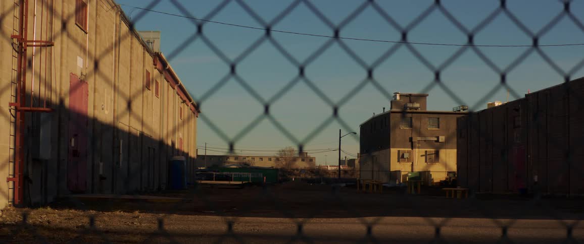 A chain link fence sits near a warehouse in a gritty city neighborhood