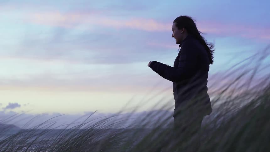 Young woman stands in grass near ocean and takes photo