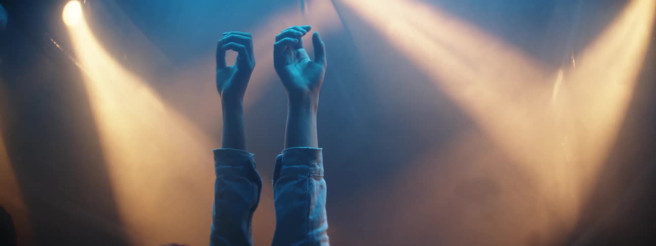 Person stands in front of reflective surfaces reaches up with one hand as lights spotlight also is reflected