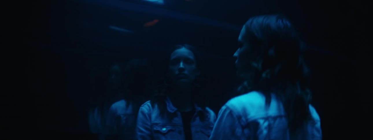 A white woman stands in front of a mirror showing her reflection as a light above flashes