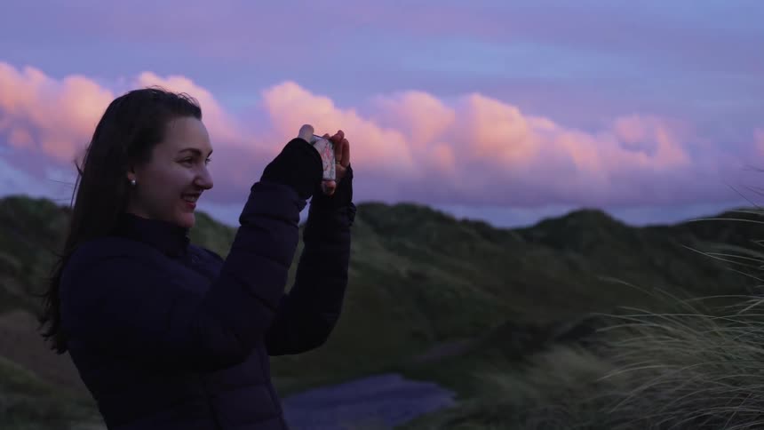 Woman standing in front of mountain range smiles and takes photo at dusk