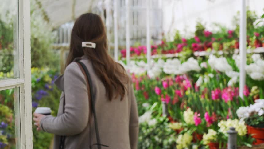 A white woman with a camera walks into a greehouse filled with plants and flowers