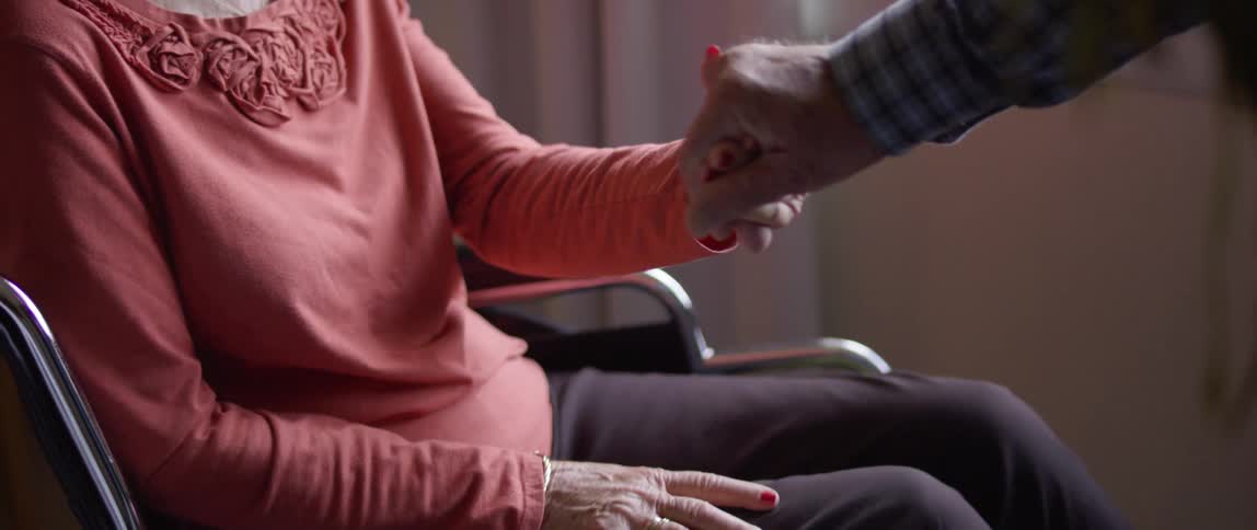 An elderly woman in a wheelchair holds hands with an elderly man