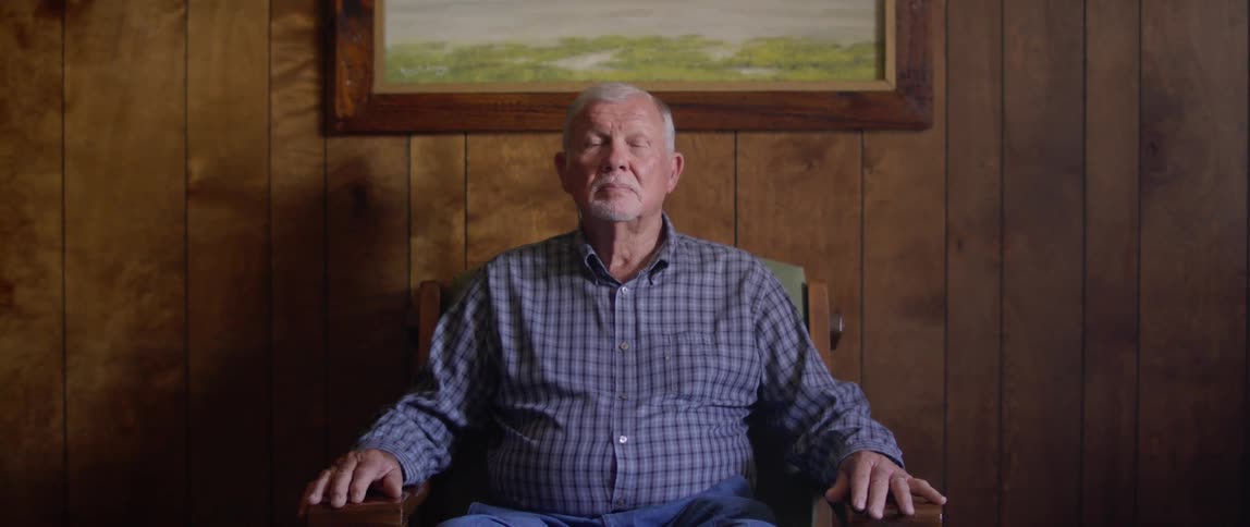 A man sits in a chair and poses in front of a painting and wood paneled wall