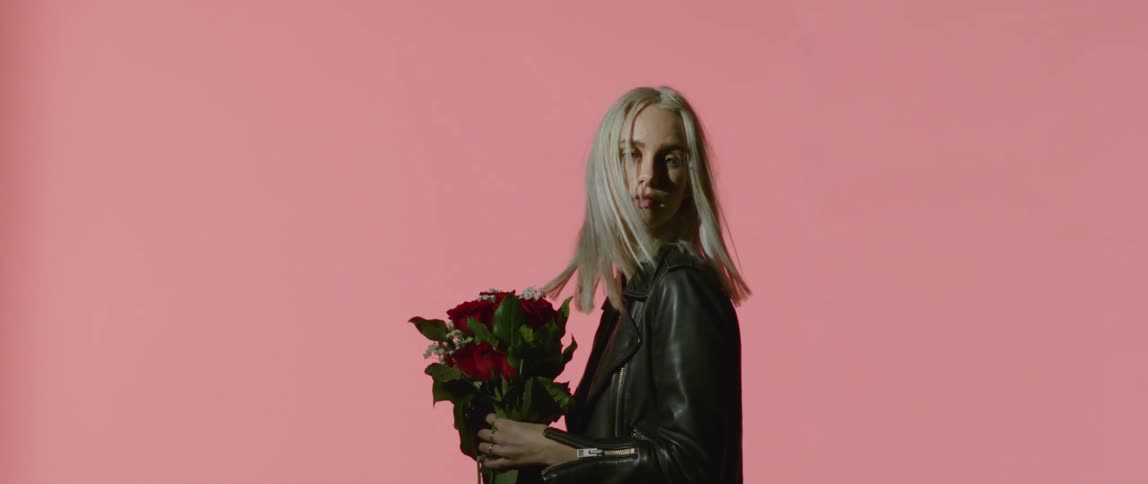 A blonde woman in a leather jacket and holding a bouquet of flowers turns her head suddenly
