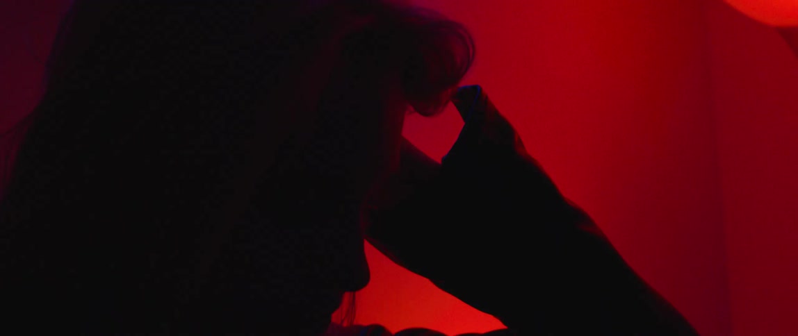 The silhouette of a woman pushing her hair back in a room with a reddish glow and a neon clock on the wall