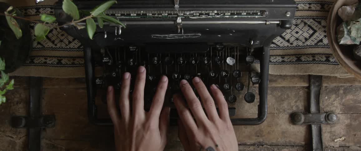 A man types on a vintage typewriter on a desk next to a plant