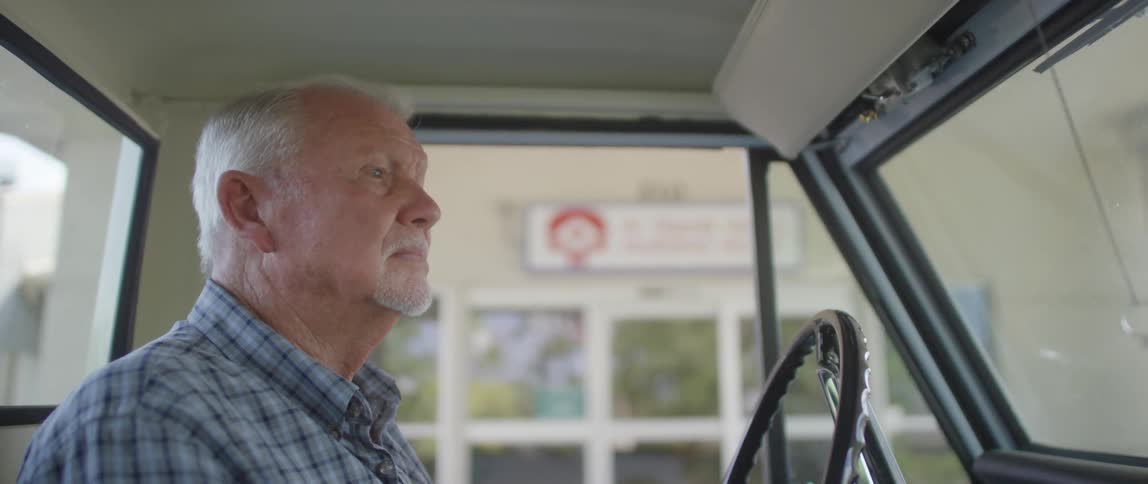 An elderly white man drives up to a store in a truck
