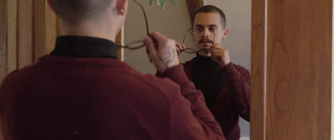 A white man stands in front of a mirror and puts his glasses on
