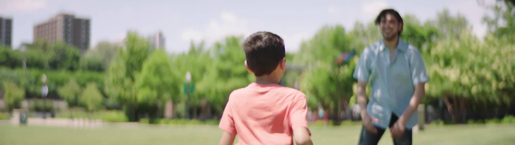 Son runs towards father who is standing near a tree and father picks son up over his head while smiling and spinning around