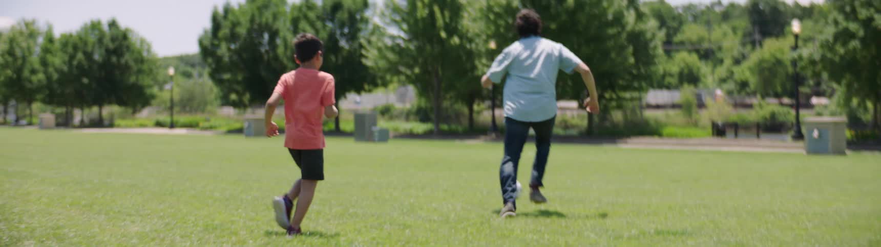 Father and son kick a soccer ball back and forth while running through a grassy area in the park