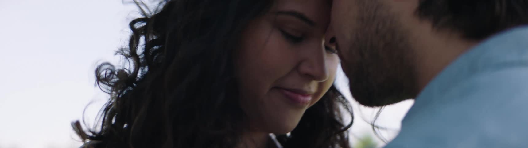 Couple lean foreheads together and clasp hands outdoors on field in park