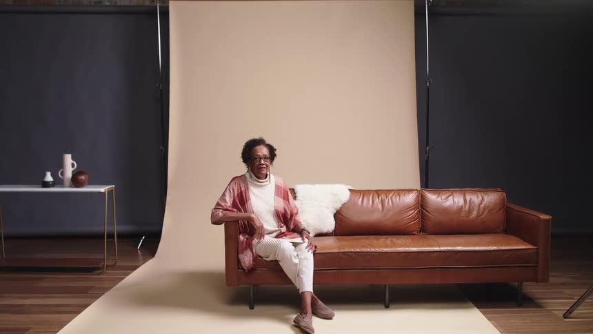 An elderly black woman poses while sitting on a couch