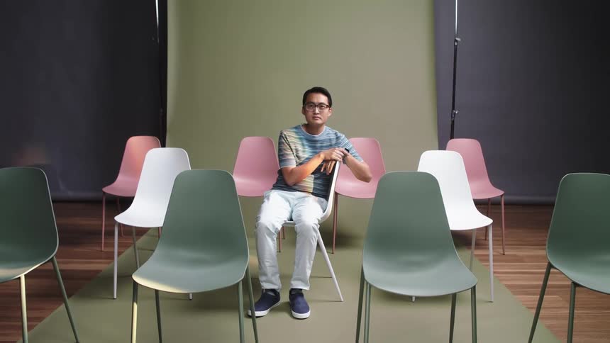 A man sits in a chair and is surrounded by colorful chairs in a photoshoot inside a studio
