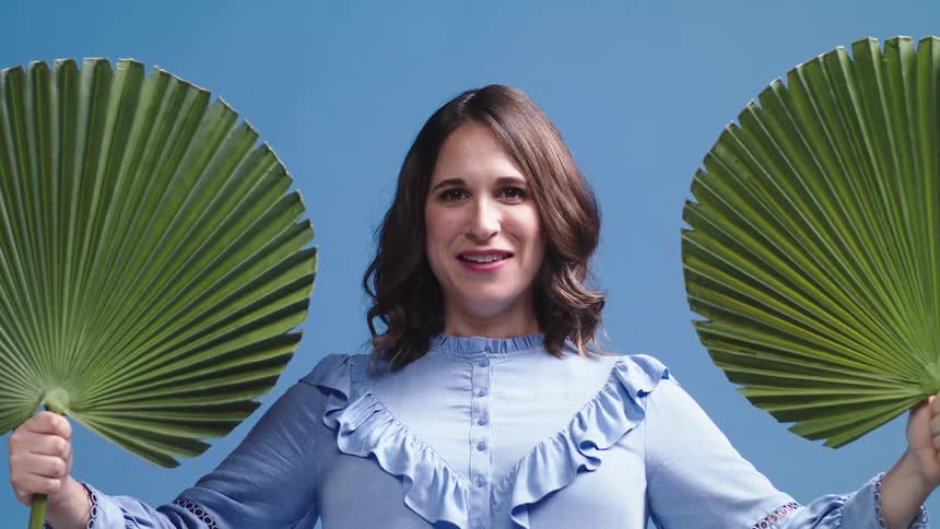 A woman in front of a back drop moves fans from in front of her face and smiles