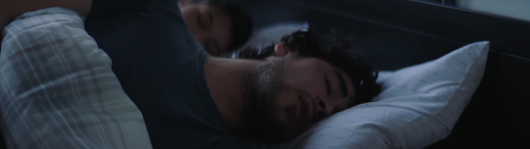 Male and female are sleeping facing the same direction in the same bed as the male starts to wake up and begins to rub his eye