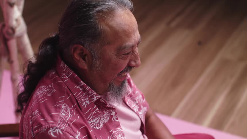 An old man sits in a chair and laughs and slaps his hands on his knees