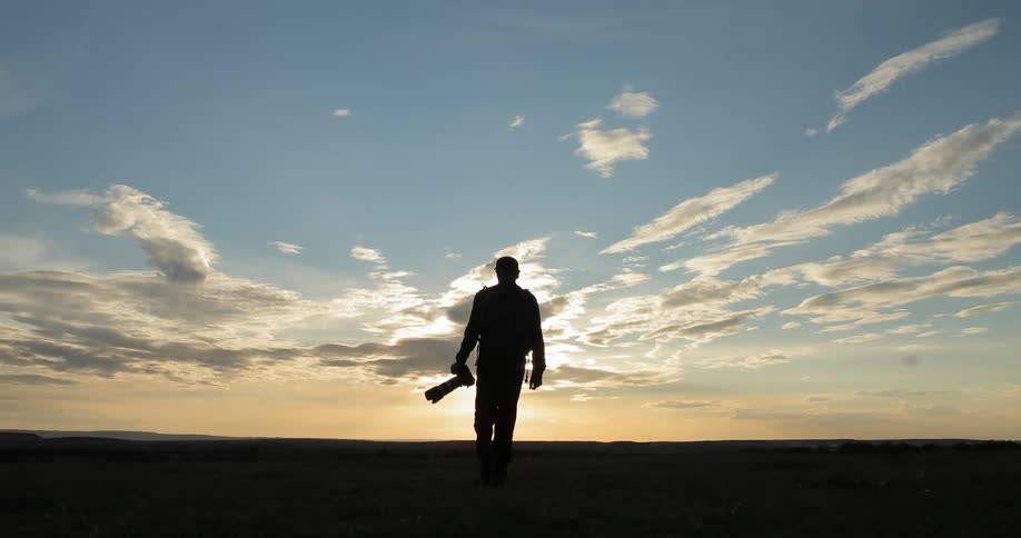 A man wearing a backpack and holding a camera takes a picture and then walks into the distance