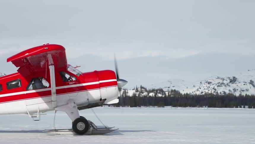 People in a red and white airplane that is on snowy terrain with trees and mountains in the background