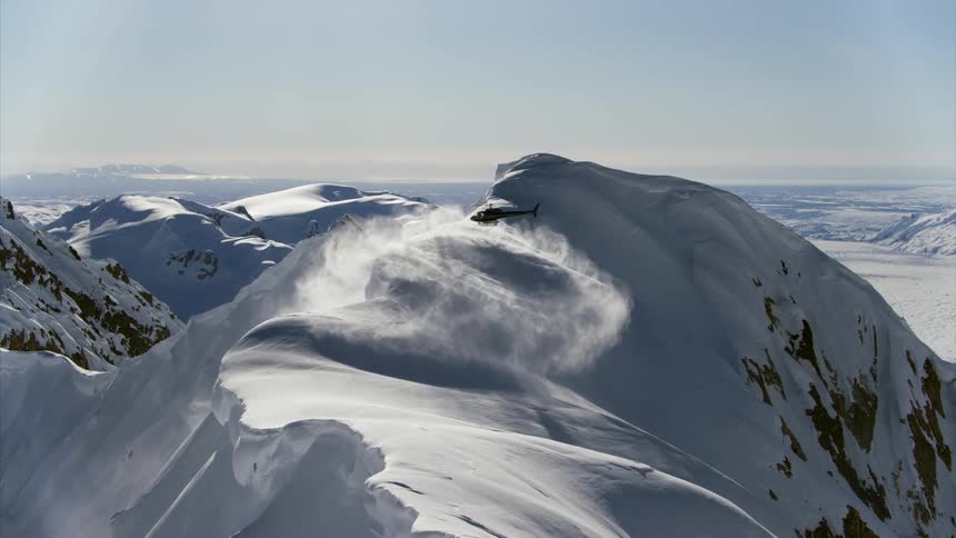 A helicopter almost lands on the peak of a snowy mountain and kicks up a cloud of snow