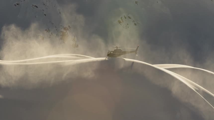 A helicopter kicks up a cloud of snow while sitting on a large drift in the mountains
