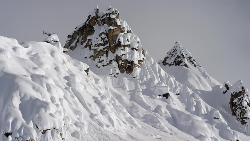 Helicopter hovers over mountain range peaks covered in snow