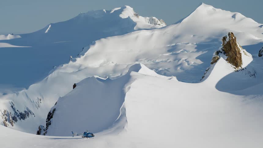 A helicopter sits on a snow drift with the engine running near a mountain