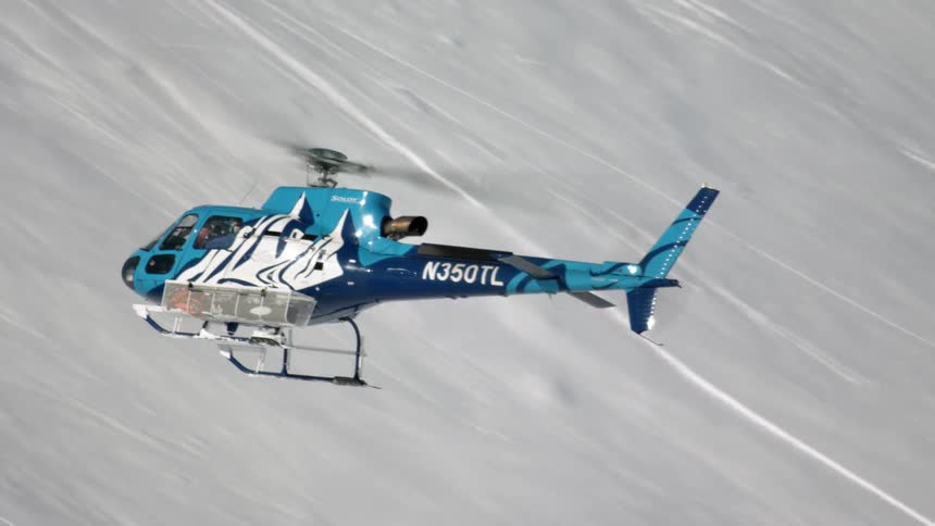 Blue helicopter with carrying case on one landing gear skims snow covered mountain slope