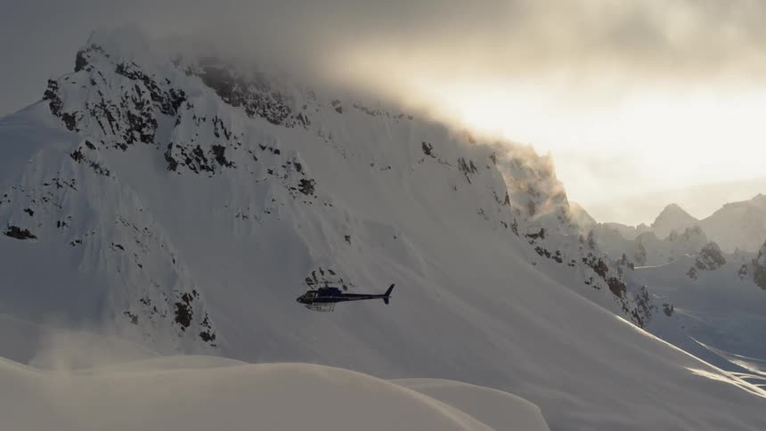 A helicopter rises up from the slope of a snow covered mountain ridge with gray clouds above