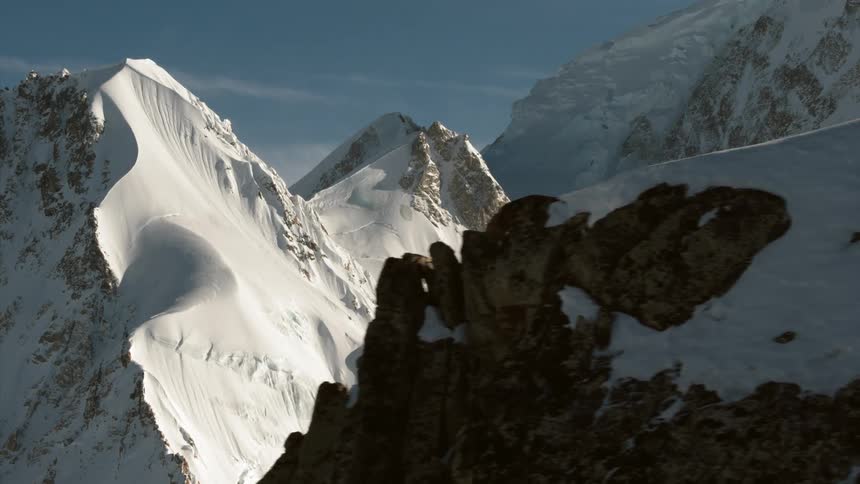 A mountain range covered in snow with a helicopter off in the distance and the sky in the background