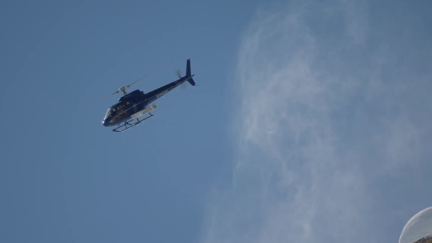 A helicopter flies over a snow covered ridge blowing snow into the bright blue sky