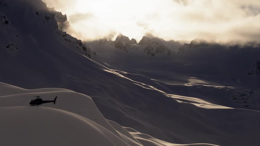 A helicopter on an extremely large mound of ice with its rotors still spinning in a mountainous snow covered arctic landscape