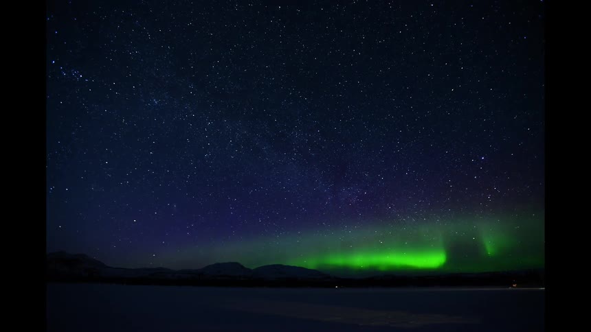 Lights flicker on plain near mountains under aurora borealis