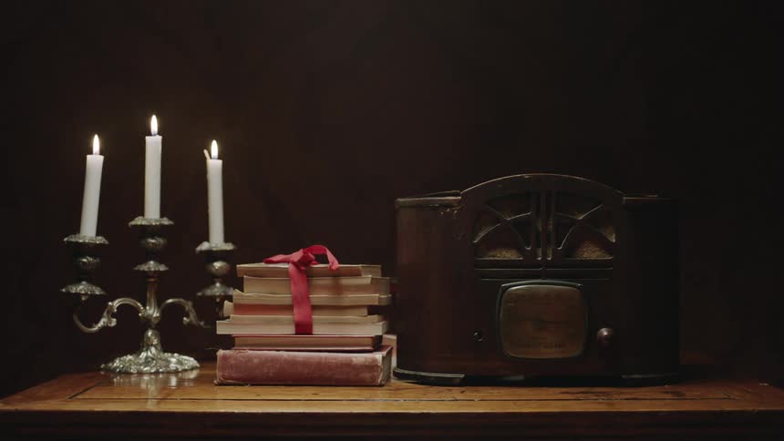 An old radio sits on a table with a stack of books and a candlestick with burning candles
