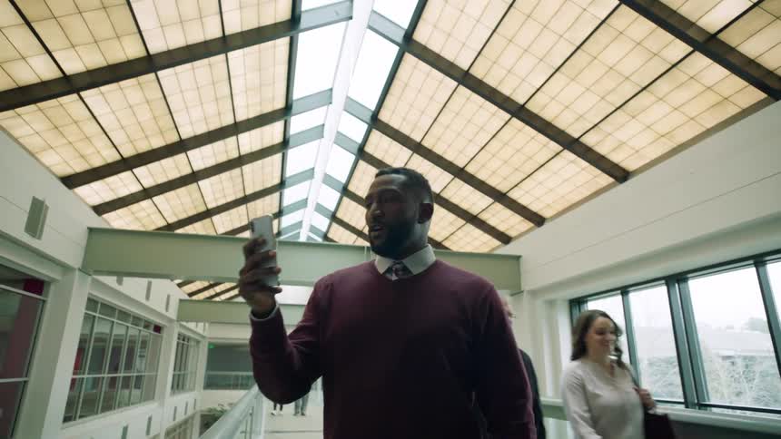 A man talks on the phone via video while walking down the hallway of a large building as people pass by