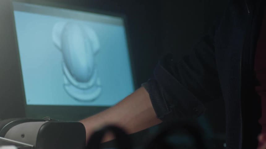 A person working at next near a monitor displaying an untextured 3d model of a virtual reality headset