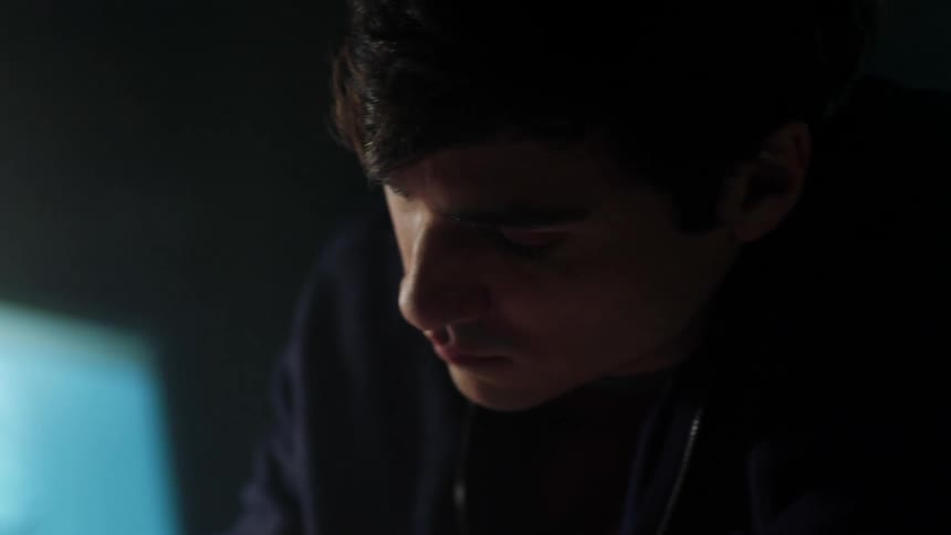 A man looks down as he sits and works with a monitor glowing beside him